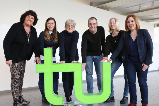 Fünf Frauen und ein Mann sehen um ein großes TU-Logo und halten es jeweils mit einer Hand fest.